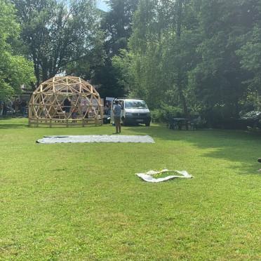 Mann sitzt in einem Stuhl auf einer Wiese vor dem wERDschätzungsrahmen und einer geodätischen Kuppel aus Holz